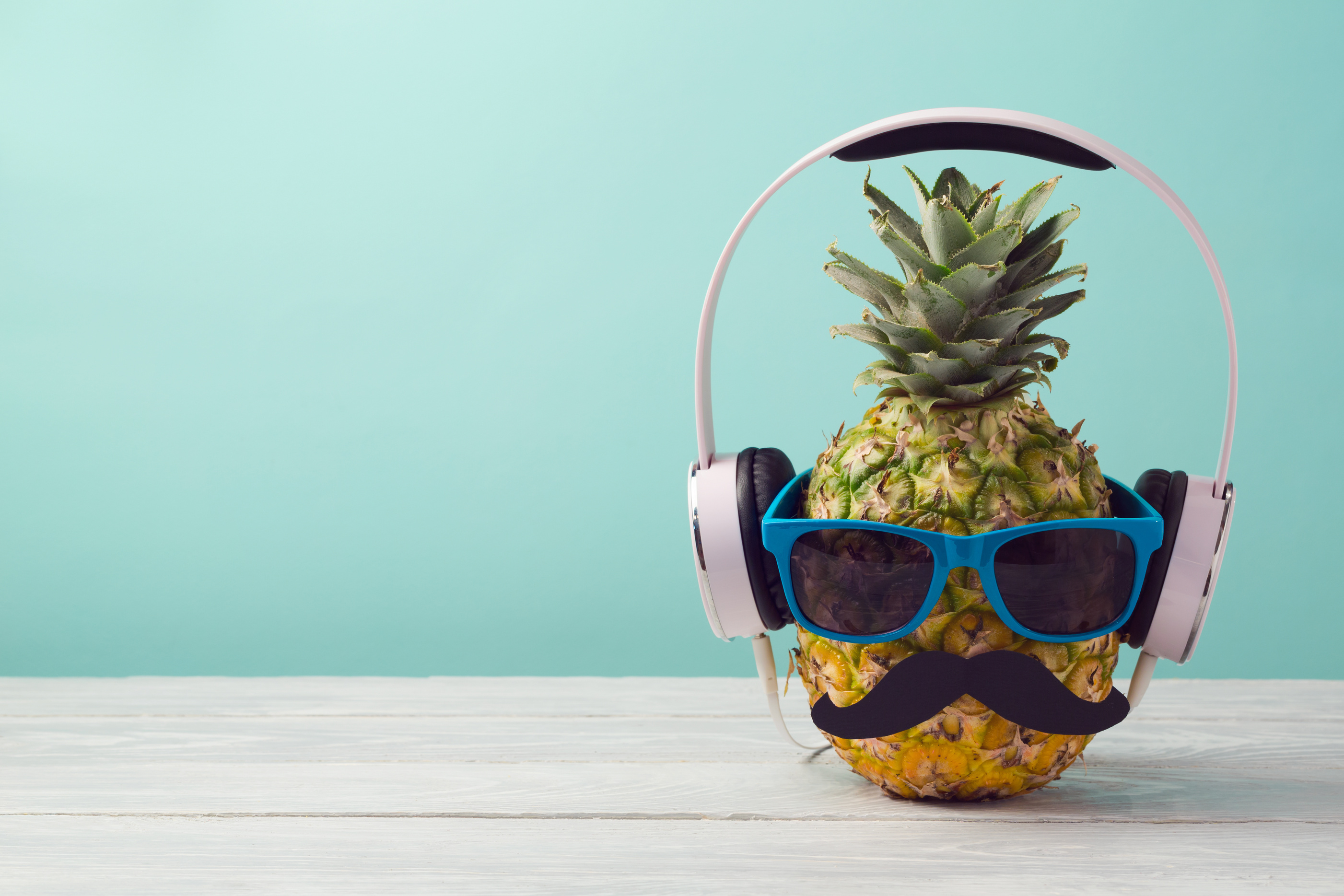Pineapple with sunglasses and headphones on wooden table over mint background. Tropical summer vacation and beach party concept.